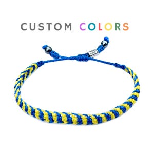 May include: Blue and yellow braided bracelet with adjustable sliding knot closure. The bracelet features a woven design with alternating blue and yellow threads. The text "CUSTOM COLORS" is displayed above the bracelet.