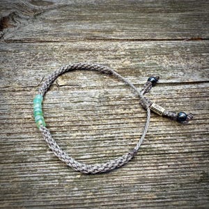 Top-down flat lay of a custom handmade rope bracelet by Rumi Sumaq on a weathered coastal deck board, showcasing the full design from above. Hand-knotted fiber cords in personalized colors frame a line of genuine blue green Peruvian Opal stones, with Hematite stone accents and a Rumi Sumaq logo bead on the adjustable drawstring tails. Handcrafted by indigenous Quechua designer Coco Paniora Salinas on Martha’s Vineyard, blending Peruvian artistry with coastal, modern unisex style.