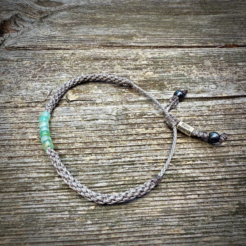 Top-down flat lay of a custom handmade rope bracelet by Rumi Sumaq on a weathered coastal deck board, showcasing the full design from above. Hand-knotted fiber cords in personalized colors frame a line of genuine blue green Peruvian Opal stones, with Hematite stone accents and a Rumi Sumaq logo bead on the adjustable drawstring tails. Handcrafted by indigenous Quechua designer Coco Paniora Salinas on Martha’s Vineyard, blending Peruvian artistry with coastal, modern unisex style.