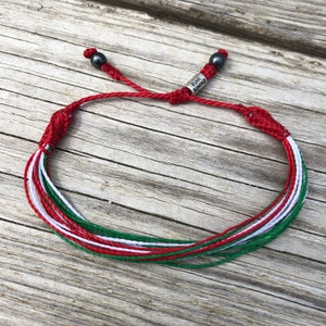 Könnte beinhalten: Geflochtenes Freundschaftsarmband in Rot, Weiß und Grün mit einem silbernen Anhänger, auf dem "Rumi Sumaq" steht.