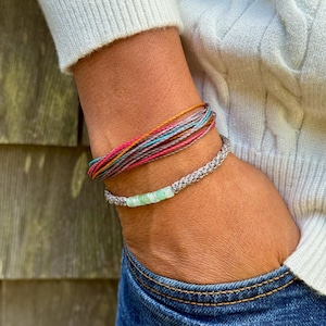 Styled image showing a stacked set of two Rumi Sumaq handmade rope bracelets layered with a surfer string bracelet on a wrist, highlighting coordinated, casual coastal style. Each custom bracelet features hand-knotted fiber cords in chosen colors, a line of blue green Peruvian Opal stones, Hematite bead accents, and a Rumi Sumaq logo bead. Handcrafted by indigenous Quechua designer Coco Paniora Salinas on Martha’s Vineyard, coastal style Opal gemstone rope bracelets for men, women, and kids.