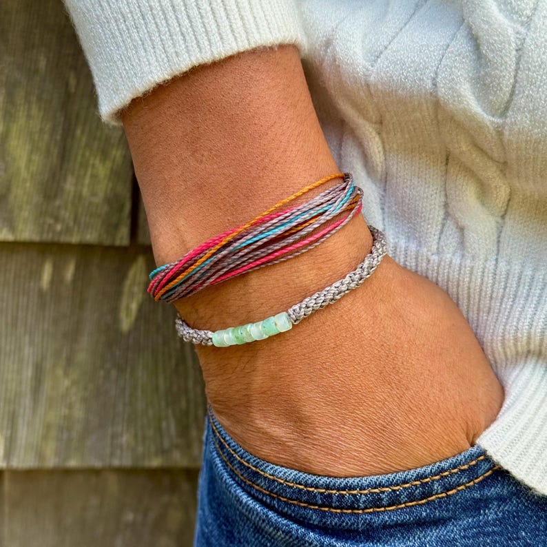 Styled image showing a stacked set of two Rumi Sumaq handmade rope bracelets layered with a surfer string bracelet on a wrist, highlighting coordinated, casual coastal style. Each custom bracelet features hand-knotted fiber cords in chosen colors, a line of blue green Peruvian Opal stones, Hematite bead accents, and a Rumi Sumaq logo bead. Handcrafted by indigenous Quechua designer Coco Paniora Salinas on Martha’s Vineyard, coastal style Opal gemstone rope bracelets for men, women, and kids.