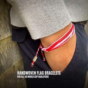 Puede incluir: Una pulsera tejida a rayas rojas, blancas y se lleva en la muñeca. La pulsera está atada con un nudo y tiene un pequeño dije de metal. El texto "HANDWOVEN FLAG BRACELETS FOR ALL 48 WORLD CUP QUALIFIERS" está en la parte inferior de la imagen.