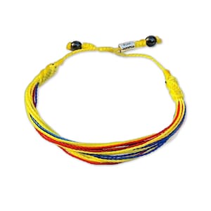 Könnte beinhalten: Ein gewebtes Armband in Gelb, Rot und Blau mit einem Schiebeknotenverschluss. Das Armband trägt den Text "RUMI SUMAQ" auf einem kleinen Metallanhänger. Das Armband besteht aus mehreren Fäden.