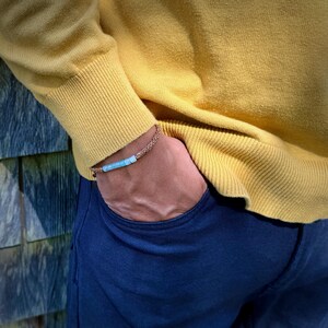 Rumi Sumaq tan rope Peruvian Opal bracelet shown on a man’s wrist, styled for everyday wear. Hand-knotted by Indigenous Quechua designer Coco on Martha’s Vineyard, this custom adjustable bracelet features seven genuine Peruvian Opal stones, Hematite bead accents, and a silver tone logo bead. A meaningful October birthstone and 24th anniversary gift for men, symbolizing creativity, balance, and calm energy in a handmade artisan design.