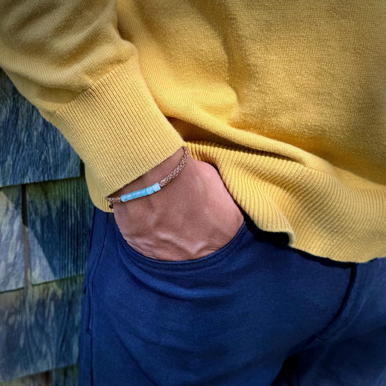 Rumi Sumaq tan rope Peruvian Opal bracelet shown on a man’s wrist, styled for everyday wear. Hand-knotted by Indigenous Quechua designer Coco on Martha’s Vineyard, this custom adjustable bracelet features seven genuine Peruvian Opal stones, Hematite bead accents, and a silver tone logo bead. A meaningful October birthstone and 24th anniversary gift for men, symbolizing creativity, balance, and calm energy in a handmade artisan design.