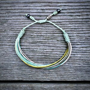 May include: A multi-colored braided bracelet with a silver charm that says "Never Give Up". The bracelet is tied with a knot and has black beads on each end.
