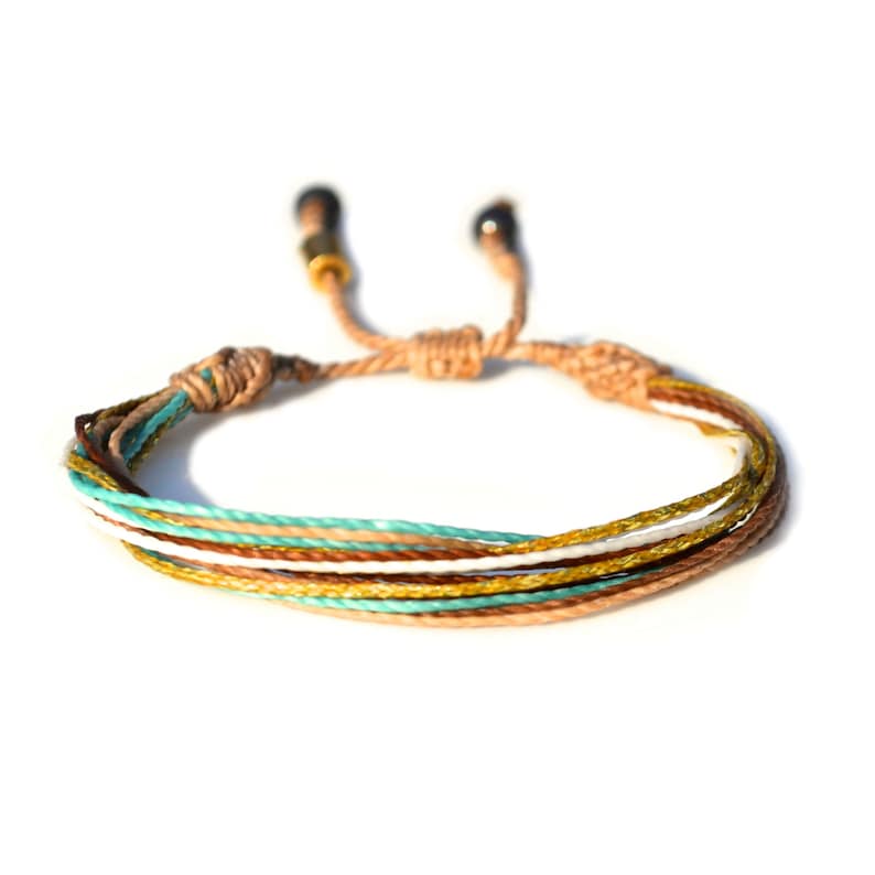 Peut inclure: Bracelet tress&eacute; multicolore avec des fils de couleur marron, blanc, or et turquoise. Le bracelet a des n&oelig;uds r&eacute;glables &agrave; chaque extr&eacute;mit&eacute;.