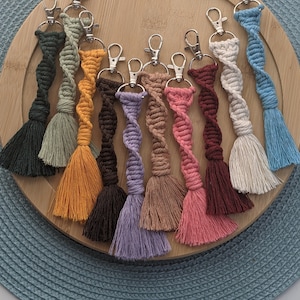 May include: A collection of 12 colorful handmade keychains with tassels. Each keychain is made with a twisted rope and features a silver key ring. The keychains are displayed on a wooden tray with a blue background.