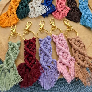 May include: A collection of 12 colorful macrame keychains with gold key rings. The keychains are made with a variety of colors, including orange, green, white, blue, pink, brown, and purple. Each keychain has a tassel at the bottom.