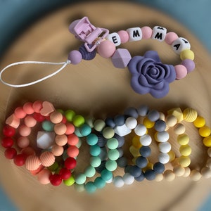 May include: A set of colorful silicone teething bracelets with different textures and shapes, including a heart, a square, and a ribbed design. The bracelets are arranged in a circle on a wooden surface. There is also a silicone teething necklace with the name "EMMA" spelled out in white letters.