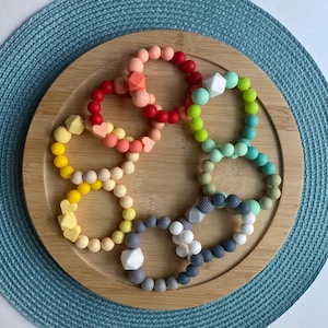 May include: A collection of colorful silicone teething bracelets in various shades of pink, red, yellow, green, and gray. Each bracelet features a unique design, including heart and geometric shapes. The bracelets are arranged on a wooden tray.