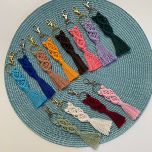 May include: A collection of 12 colorful macrame keychains with tassels. Each keychain features a unique color and pattern, including blue, green, pink, red, and white.