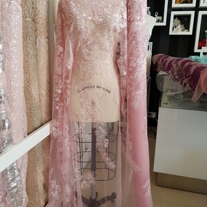 May include: Pink tulle fabric with delicate white floral embroidery and sequins. The fabric is draped over a dress form.