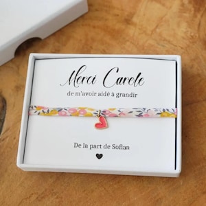 Peut inclure: Une boîte cadeau blanche avec une carte portant l'inscription "Merci Carole" et un bracelet floral avec un charm en forme de cœur rouge. La carte porte également l'inscription "De la part de Sofian" et un cœur noir.