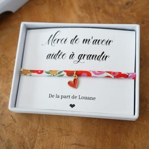 Peut inclure: Un coffret blanc contient un bracelet avec un bracelet à motifs floraux et un charm en forme de cœur rouge. La carte à l'intérieur de la boîte porte du texte en français et un petit cœur noir. Le bracelet est présenté sur une surface en bois.