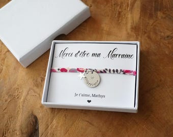 May include: A fabric bracelet with a floral pattern in pink, white, and grey, featuring a silver star charm and a round silver disc engraved with a heart and the word marraine. The bracelet is displayed on a white card with the text Merci d'être ma Marraine and Je t'aime, Mathys, inside a white gift box.