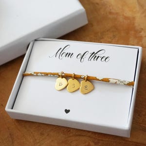 Peut inclure: Bracelet à breloques en forme de cœur doré avec les lettres B, S et E, présenté dans un coffret cadeau blanc. La carte à l'intérieur de la boîte indique "Mom of three". Le bracelet est sur un bracelet à motifs.