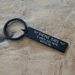 Peut inclure: Porte-clés rectangulaire noir avec un anneau noir. Le porte-clés porte le texte blanc "DRIVE SAFE I NEED YOU HERE WITH ME" avec des symboles de cœur. Le porte-clés est sur une surface en bois brun clair.