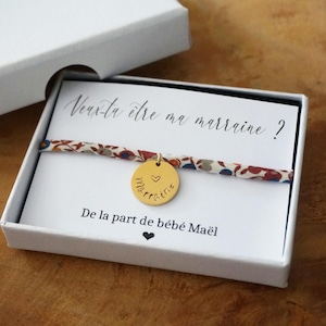 Peut inclure: Un bracelet en tissu à motif floral avec une breloque ronde dorée gravée d'un cœur et du mot marraine. Le bracelet est présenté dans une boîte blanche sur une carte avec le texte Veux-tu être ma marraine? De la part de bébé Maël.