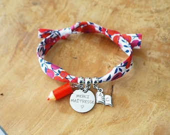 Puede incluir: Una pulsera de tela con un estampado floral rojo, rosa y blanco. La pulsera tiene un dije de lápiz rojo, un dije plateado con la inscripción "MERCI MAÎTRESSE" y un dije de libro. Sobre una superficie de madera.