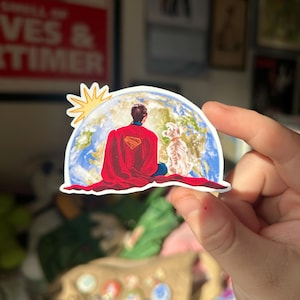 May include: A sticker featuring a superhero in a red cape, seated in a meditative pose on a globe, with a dog beside him. The sun shines above. The sticker has a white border and is held by a hand.