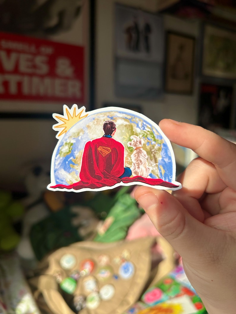 May include: A sticker featuring a superhero in a red cape, seated in a meditative pose on a globe, with a dog beside him. The sun shines above. The sticker has a white border and is held by a hand.