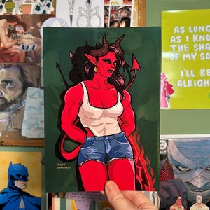 May include: A print featuring a red-skinned character with horns, a white tank top, and denim shorts. The artwork includes a tail and a pitchfork. The artist's name, "THEFT SHRJEERY", is visible. The background is green.