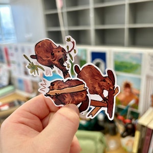 May include: Three brown and white beaver stickers on a white background. The beavers are depicted in different poses, one is holding a branch, one is gnawing on a branch, and one is sitting on a branch.