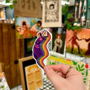 May include: A sticker featuring a cartoon illustration of a person wearing a purple robe and a pointed hat, holding a wand and standing in front of a rainbow.
