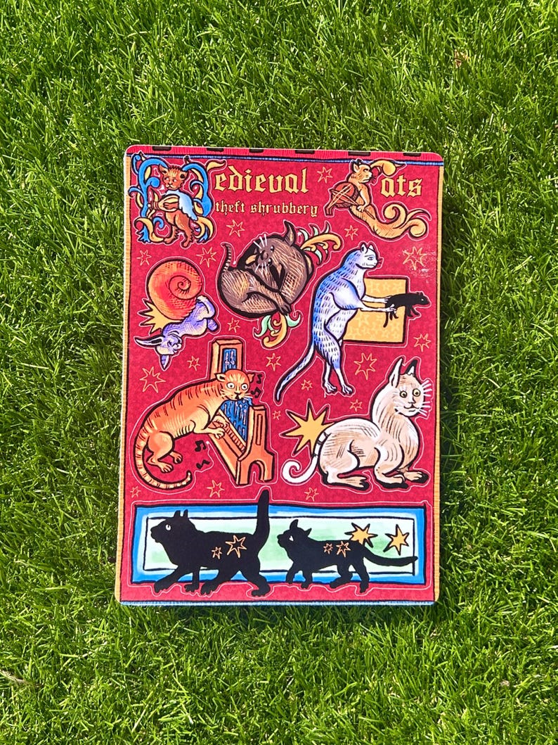 Puede incluir: Un cuaderno rectangular rojo con las palabras "Medieval Cats" y "theft shrubbery" impresas en la portada. La portada presenta varias ilustraciones de gatos de dibujos animados al estilo medieval, con estrellas y elementos decorativos.