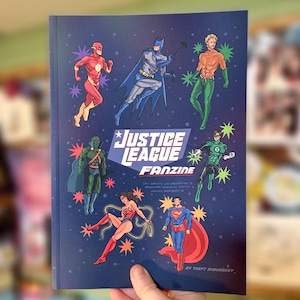 May include: A Justice League Fanzine held in a hand. The cover shows illustrations of the Flash, Batman, Aquaman, Martian Manhunter, Wonder Woman, Superman, and Green Lantern against a dark blue background with starbursts. The title "Justice League Fanzine" is displayed.