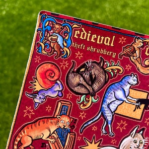 Puede incluir: Una hoja de pegatinas con un fondo burdeos que presenta ilustraciones de estilo medieval de gatos y otros animales. El texto "Medieval theft shrubbery" est&aacute; en la parte superior. Las ilustraciones son en tonos naranja, azul, marr&oacute;n y morado.