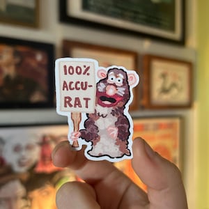 May include: A sticker depicting a cartoon rat holding a sign that reads "100% ACCU-RAT." The rat is coloured brown, white, and pink, with a surprised expression. The sticker is held in a person's hand, with framed artwork in the background.