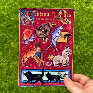 Puede incluir: Una pegatina rectangular con un fondo rojo que presenta ilustraciones de gatos y otras criaturas de estilo medieval. El texto "Medieval Cats" y "theft shrubbery" es visible. La pegatina tiene un borde azul y rojo y est&aacute; sujeta por una mano.