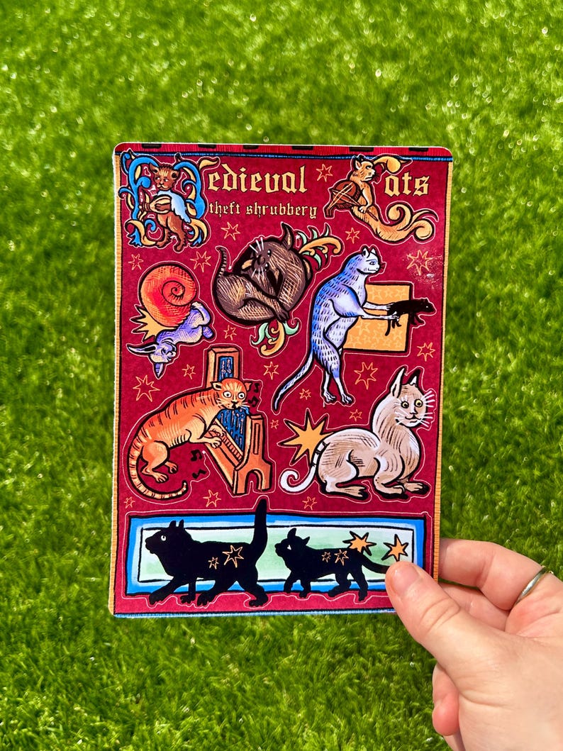 Puede incluir: Una pegatina rectangular con un fondo rojo que presenta ilustraciones de gatos y otras criaturas de estilo medieval. El texto "Medieval Cats" y "theft shrubbery" es visible. La pegatina tiene un borde azul y rojo y est&aacute; sujeta por una mano.