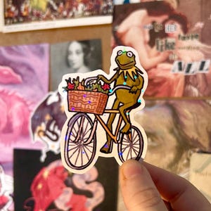May include: A die-cut sticker featuring a green frog character riding an orange bicycle. The bicycle has a brown wicker basket attached to the handlebars, filled with colourful flowers. The sticker has a holographic finish with small star accents.