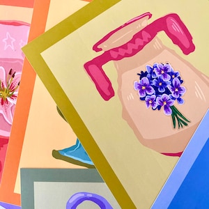 Puede incluir: Una pila de tarjetas coloridas con ilustraciones de varios objetos, incluyendo una jarra con un ramo de flores moradas, una jarra azul y una jarra morada. Las tarjetas tienen un borde dorado, naranja y gris.