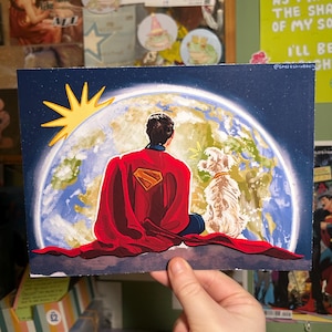 May include: A print featuring Superman in a red cape with the Superman logo, looking at Earth with a dog. The sun is depicted in yellow. The background is a dark blue sky. The print is held by a hand.