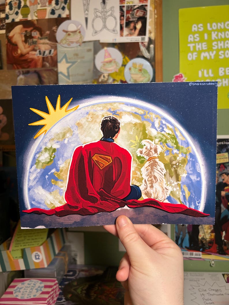May include: A print featuring Superman in a red cape with the Superman logo, looking at Earth with a dog. The sun is depicted in yellow. The background is a dark blue sky. The print is held by a hand.