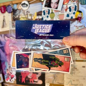 May include: A clear plastic bag containing a Justice League sticker pack by Theft Strawberry. The header card features a dark blue starry background with white text. Inside are rectangular stickers depicting comic book characters, including a green figure in a cape and a red-clad character, each with a small numbered box in the corner.