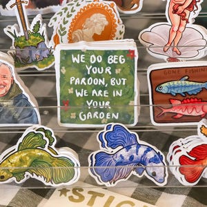May include: A collection of colourful stickers featuring various designs, including a person with a sword, a fish, a woman in a dress, and a fish tank. The stickers have a whimsical and playful aesthetic. One sticker reads "We do beg your pardon, but we are in your garden."