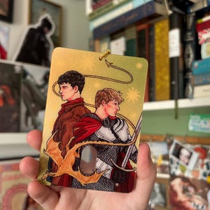 May include: A rectangular acrylic charm featuring a glittery gold background with a printed illustration of two figures standing back-to-back. One figure wears a brown jacket, while the other wears silver chainmail armor and a red cape with a sword.