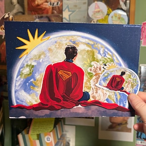 May include: A Superman-themed art print depicts Superman with his back to the viewer, gazing at Earth. The artwork features a red cape, the Superman logo, and a stylized sun. A smaller, similar image is held in a hand.