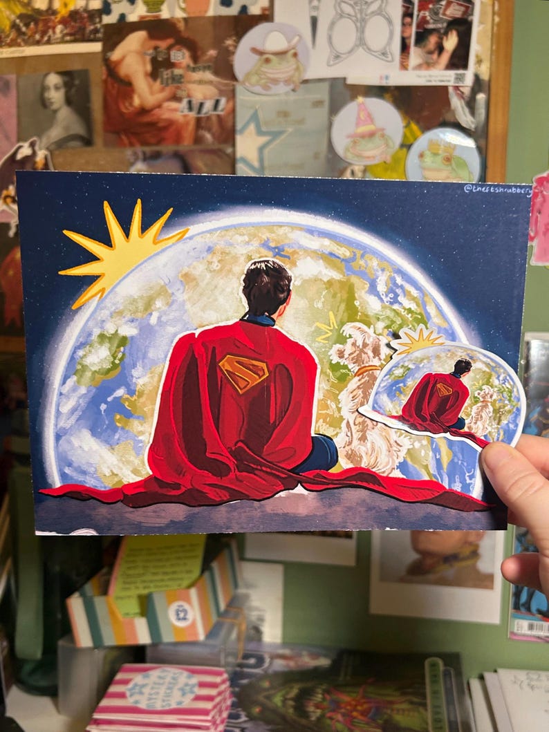 May include: A Superman-themed art print depicts Superman with his back to the viewer, gazing at Earth. The artwork features a red cape, the Superman logo, and a stylized sun. A smaller, similar image is held in a hand.
