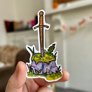 May include: A sticker of a golden sword stuck in a purple rock with green moss and a white flower.