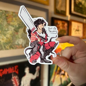 May include: A sticker featuring a warrior woman with a large sword, dressed in red and black armor. The sticker has the text "Theft Shrubbery" and is held by a hand.