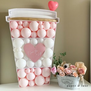 K&ouml;nnte beinhalten: Ein wei&szlig;er Pappbecher, gef&uuml;llt mit rosa und wei&szlig;en Luftballons. Die Ballons sind in einem Muster angeordnet, das einem Kaffeebecher &auml;hnelt. Ein rosa Herzballon befindet sich oben auf dem Becher. Ein rosa Herz mit dem Text "I Love You a Latte" befindet sich in der Mitte des Bechers. Im Vordergrund steht ein kleiner Eimer aus Metall mit Blumen. Der Eimer tr&auml;gt den Text "Flowers & Garden paper & luxe decor" auf der Seite.