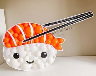 Puede incluir: Una escultura de globos de un rollo de sushi con palillos. El sushi está hecho de globos blancos con rayas naranjas y blancas para el salmón y ojos negros y una sonrisa. Los palillos son negros y están hechos de dos globos largos.