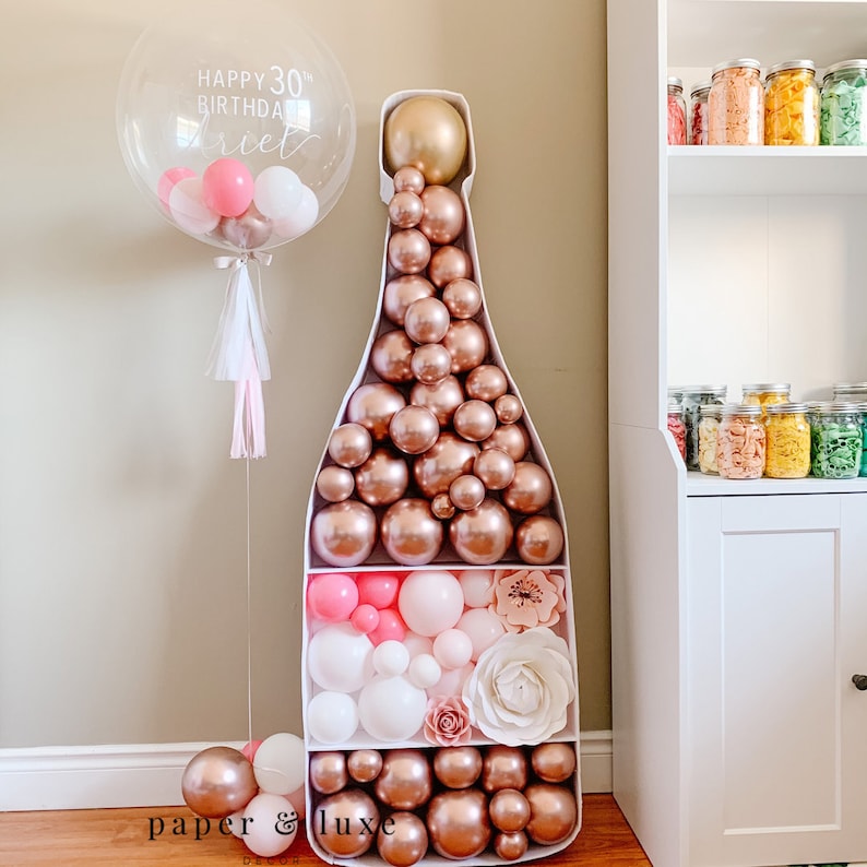 May include: A white champagne bottle shaped balloon sculpture filled with rose gold and white balloons. The sculpture is decorated with pink and white flowers. The text "Happy 30th Birthday" is written on a clear balloon.